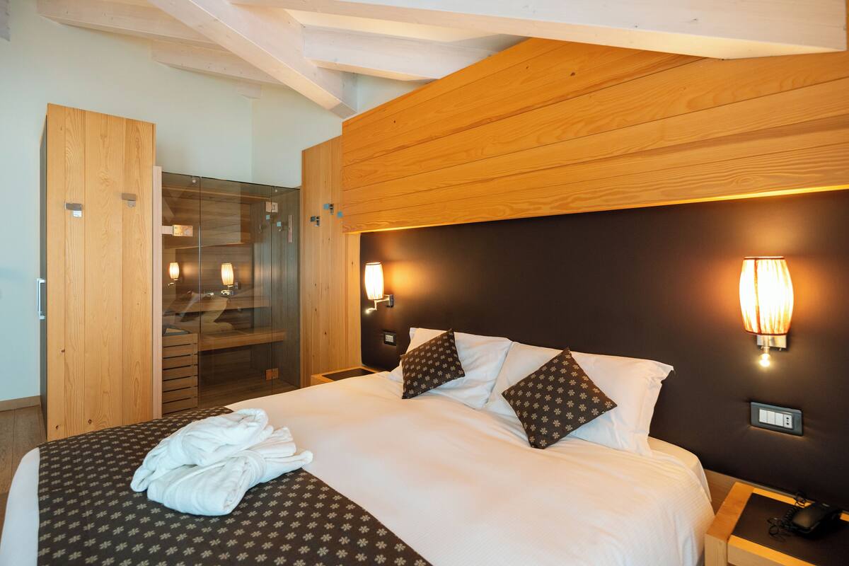 A hotel room with a bed, a wooden headboard, a glass shower enclosure, and a nightstand with a phone.
