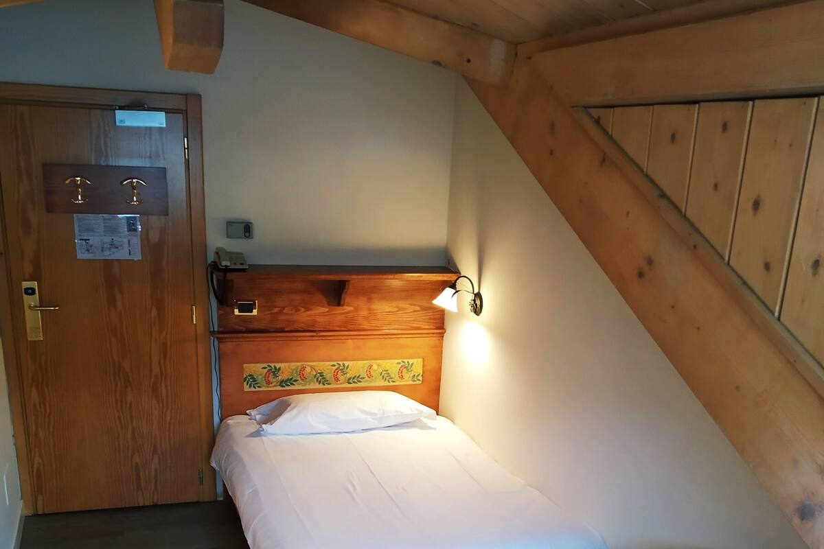 A small, cozy room with a wooden ceiling, a bed with a headboard, a bedside table with a lamp, and a wooden door.
