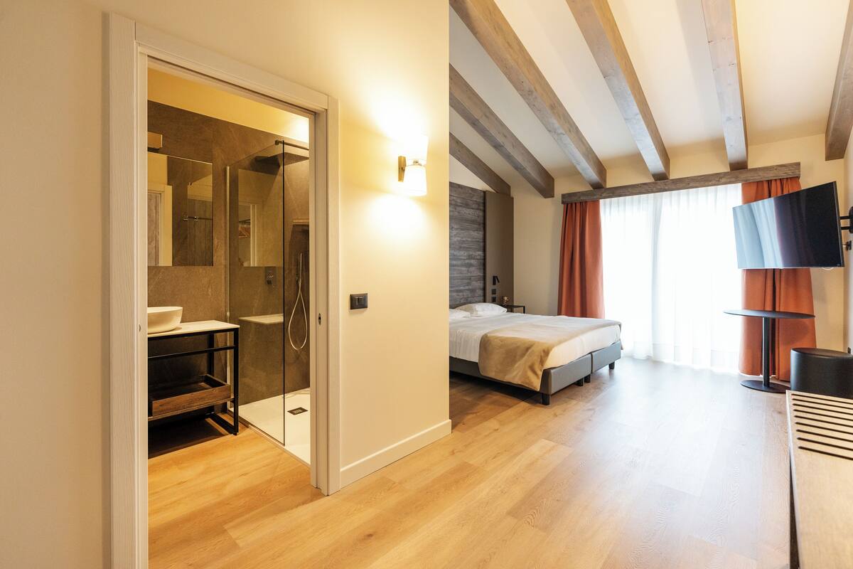 A modern hotel room with a wooden floor, a bed, a TV, and a bathroom with a glass shower enclosure.
