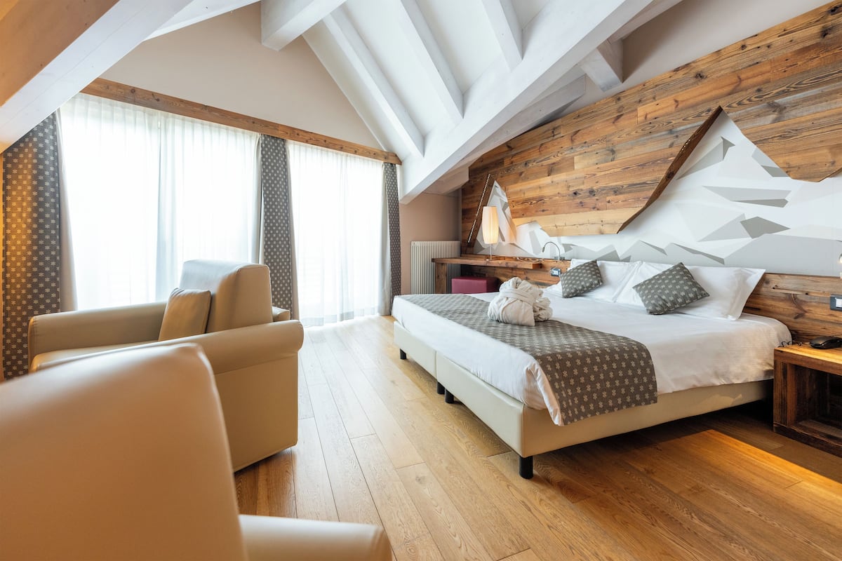 A modern hotel room with a wooden ceiling, a large bed with patterned bedding, a seating area with a sofa, and a view through a window with curtains.