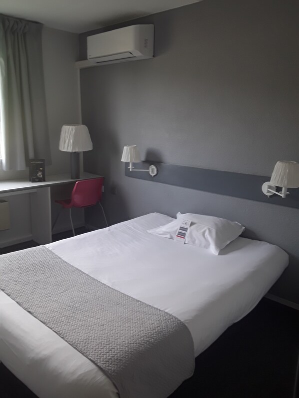 Quarto de hotel com cama, escrivaninha, cadeira e ar-condicionado na parede.