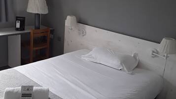 Ein ordentlich bezogenes Bett mit weißen Leinentüchern, ein Nachttisch mit Lampe sowie ein kleiner Schreibtisch mit Buch und Fernbedienung.