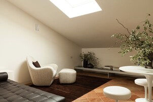 A modern living room with a high ceiling, a skylight, a black leather sofa, a white armchair, a small ottoman, a white table, and a potted plant.