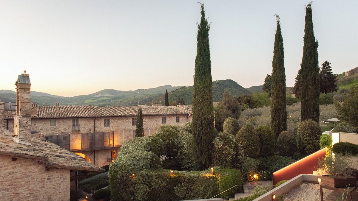 Un edificio histórico con un patio, altos cipreses y montañas al fondo.