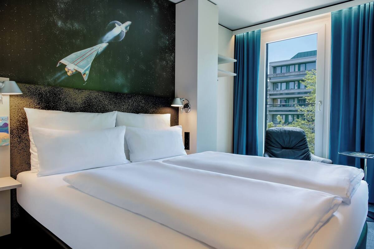 Camera d'albergo con un letto grande, un comodino, una lampada a parete, una finestra con tende e vista su un edificio.