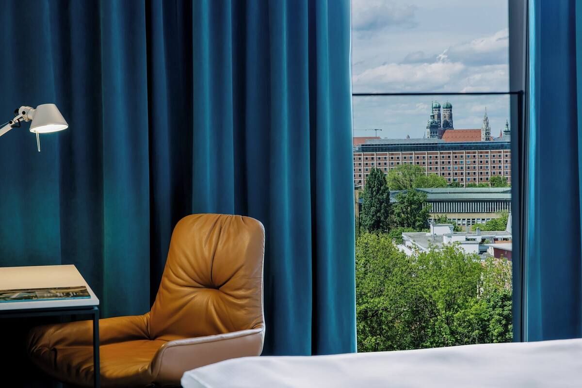 Habitación de hotel con vistas a un paisaje urbano, un sillón de cuero, un escritorio con lámpara y una cama.