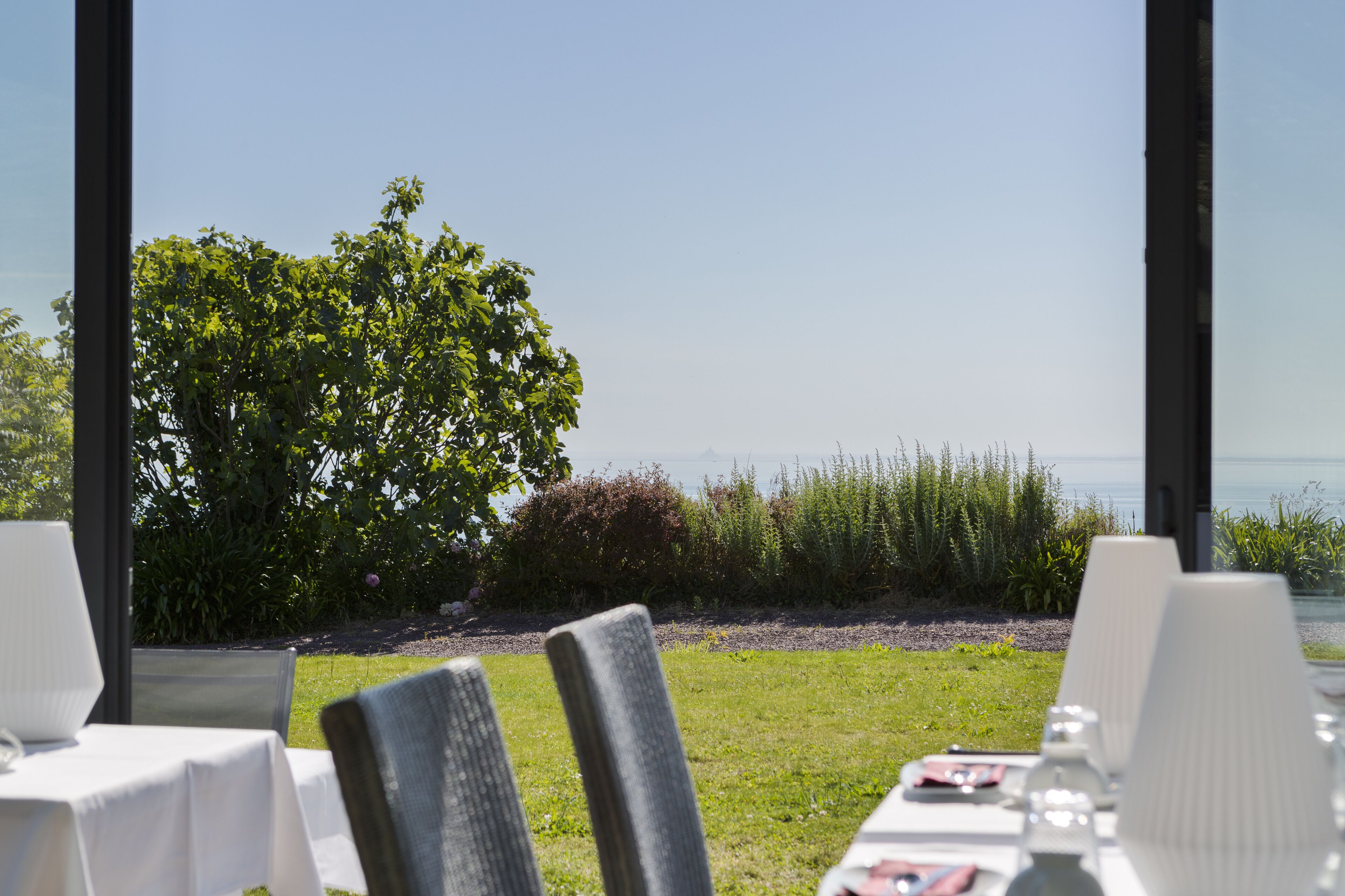 An outdoor dining area with tables and chairs, a view of the sea, and greenery in the background.