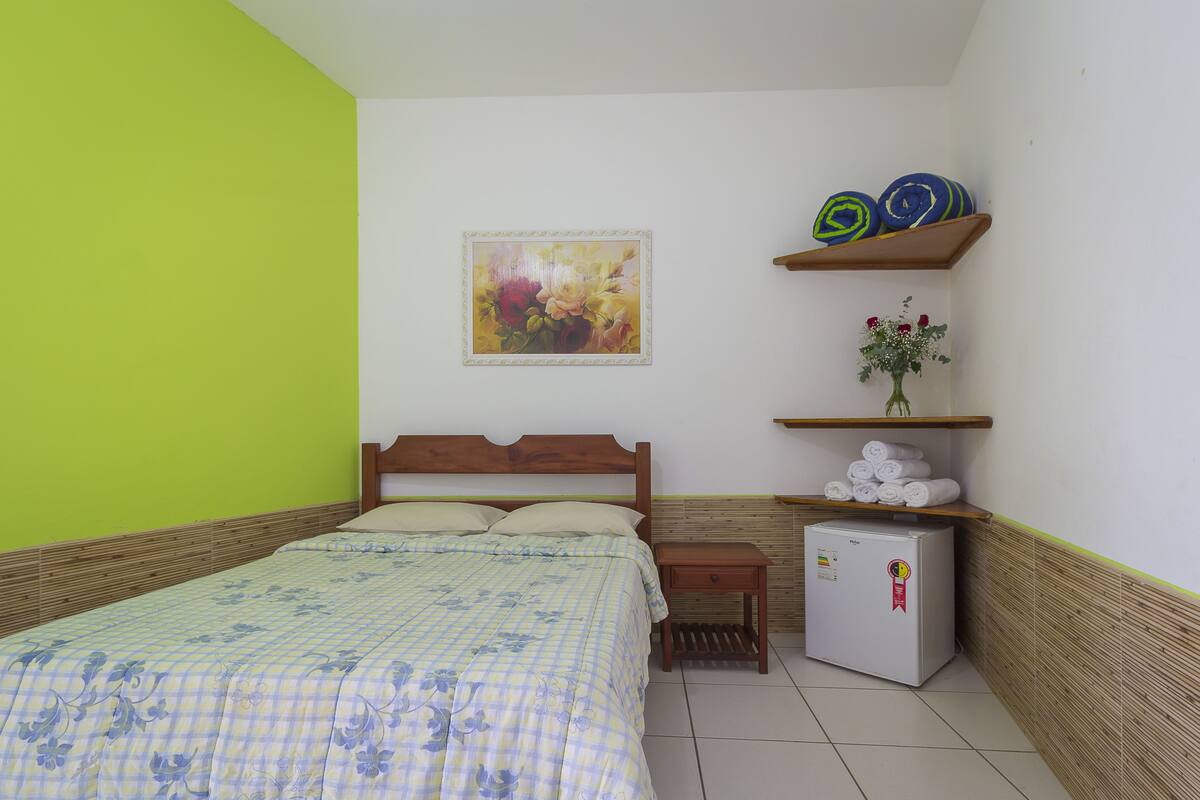 A bedroom with a bed, a small bedside table, a wall-mounted shelf with towels and a vase of flowers, and a refrigerator.