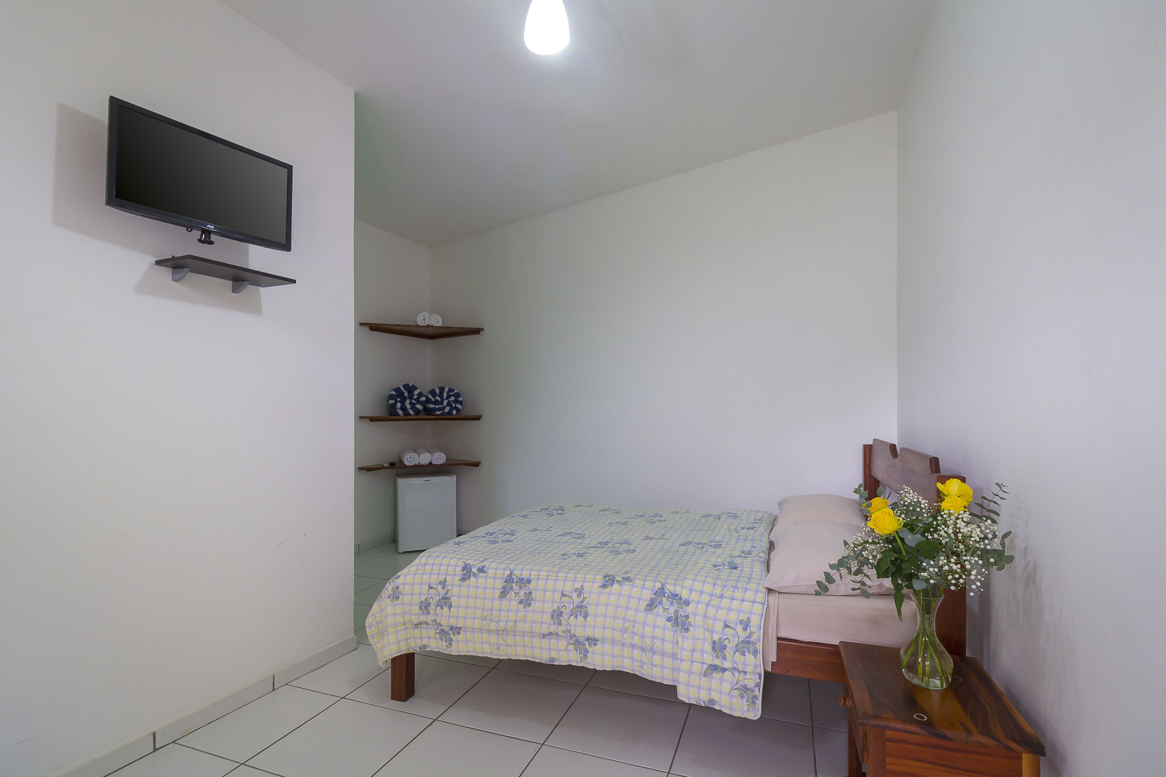 A bedroom with a bed, a TV mounted on the wall, a small shelf with decorative items, and a vase with flowers on a bedside table.