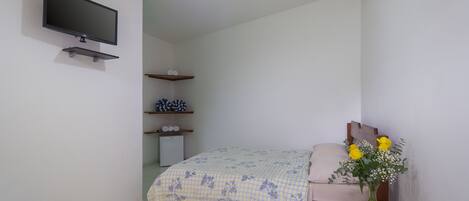 A bedroom with a bed, a TV mounted on the wall, a small shelf with decorative items, and a vase with flowers on a bedside table.