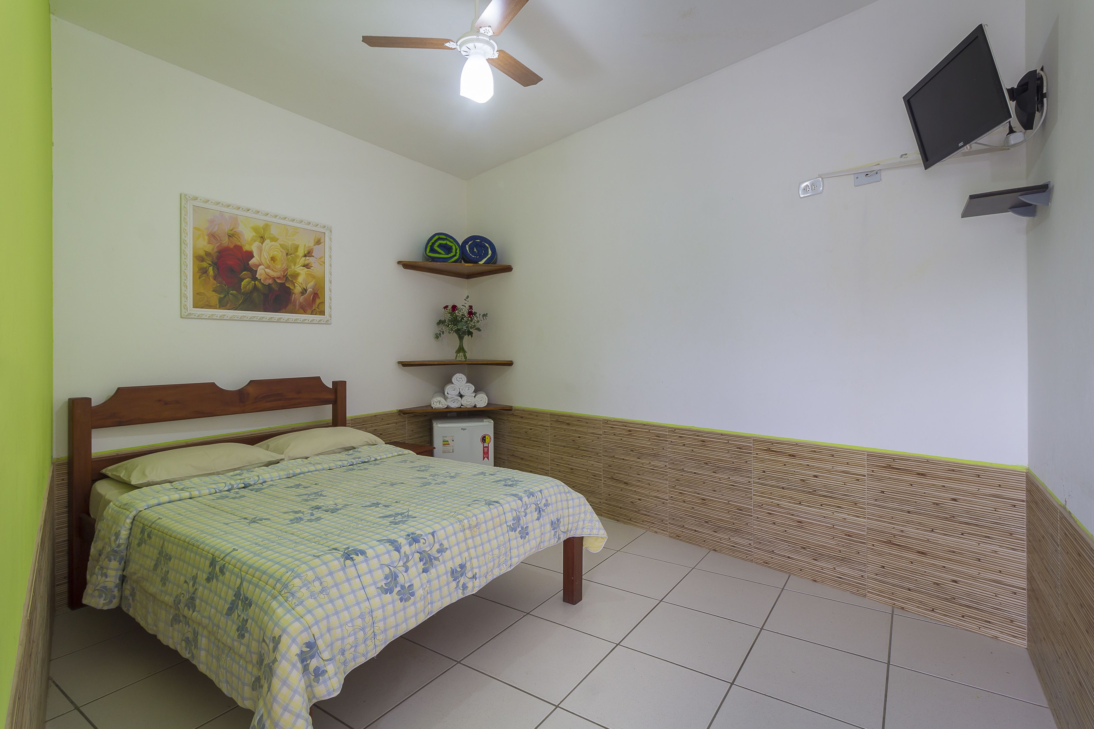 A bedroom with a bed, a ceiling fan, a mounted TV, and a wall-mounted shelf with decorative items.