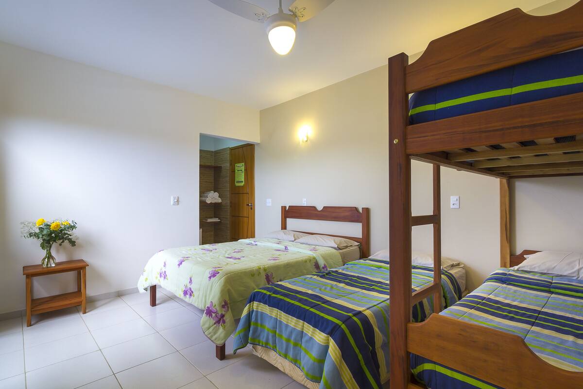 A room with two single beds, a wooden nightstand with a vase of flowers, and a ceiling fan.