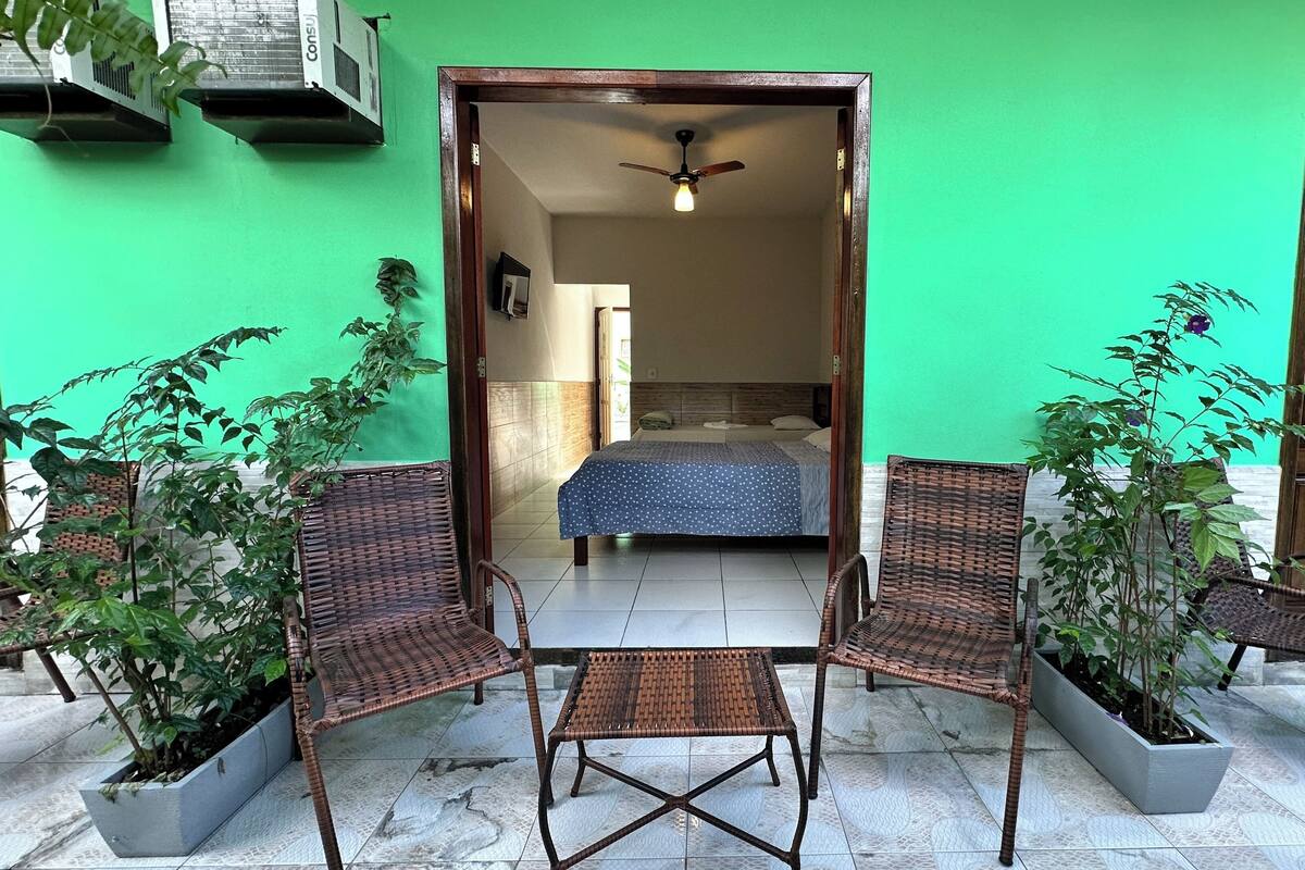 A room with a bed, two wicker chairs, a small table, and potted plants.
