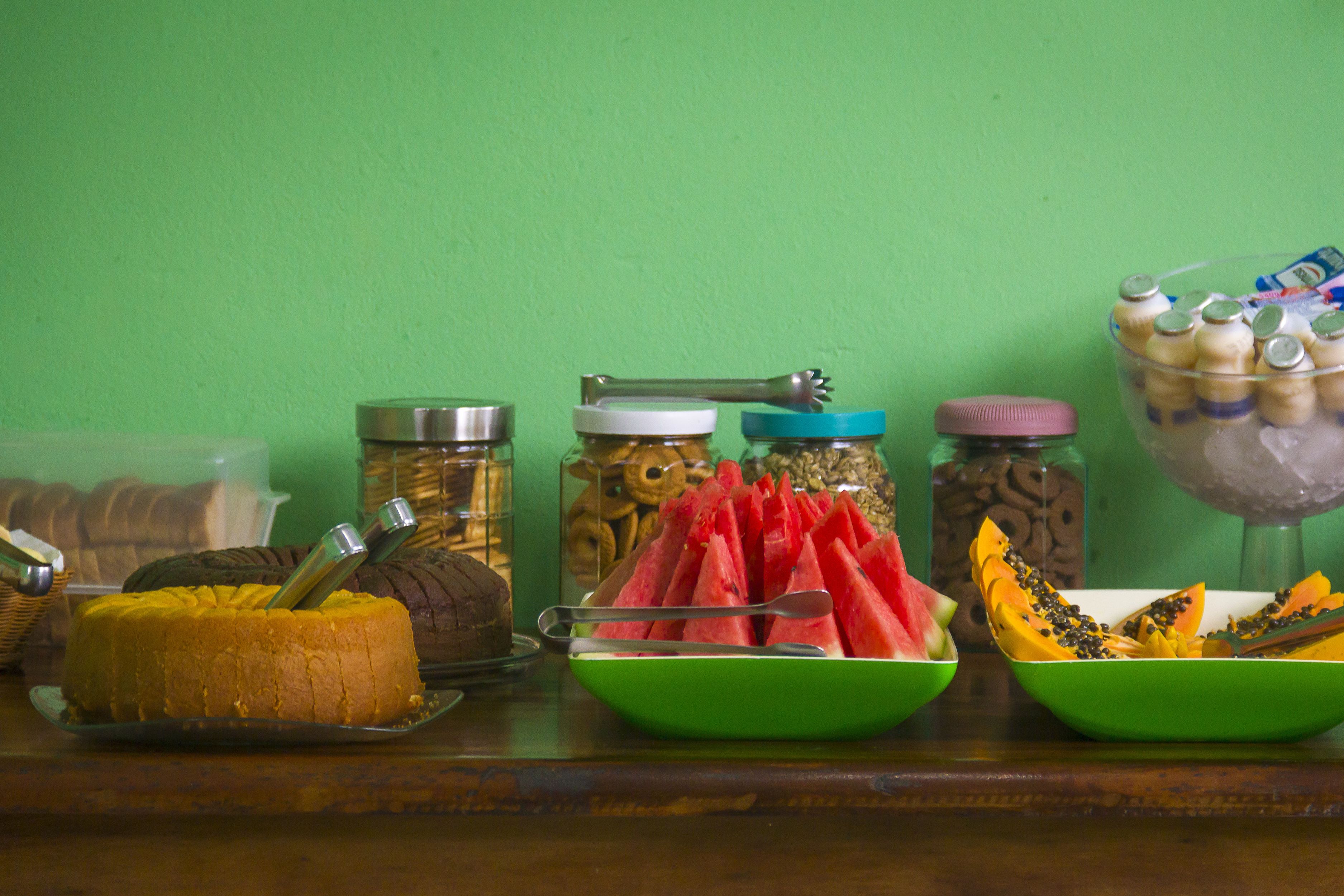 A variety of food items including a cake, watermelon, and bowls of fruit on a table against a green wall.