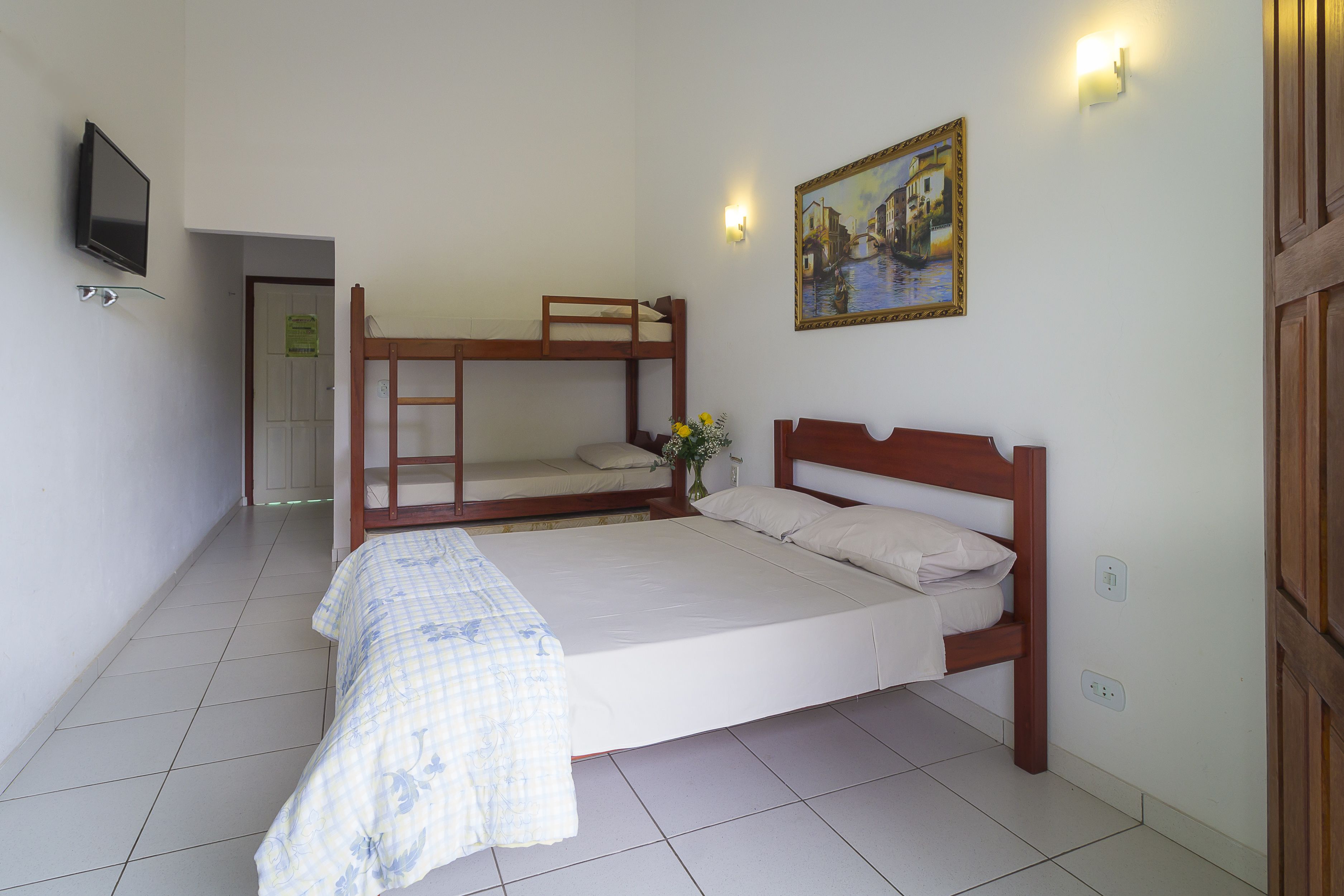 A room with a bunk bed, a single bed, a TV mounted on the wall, and a painting above the single bed.