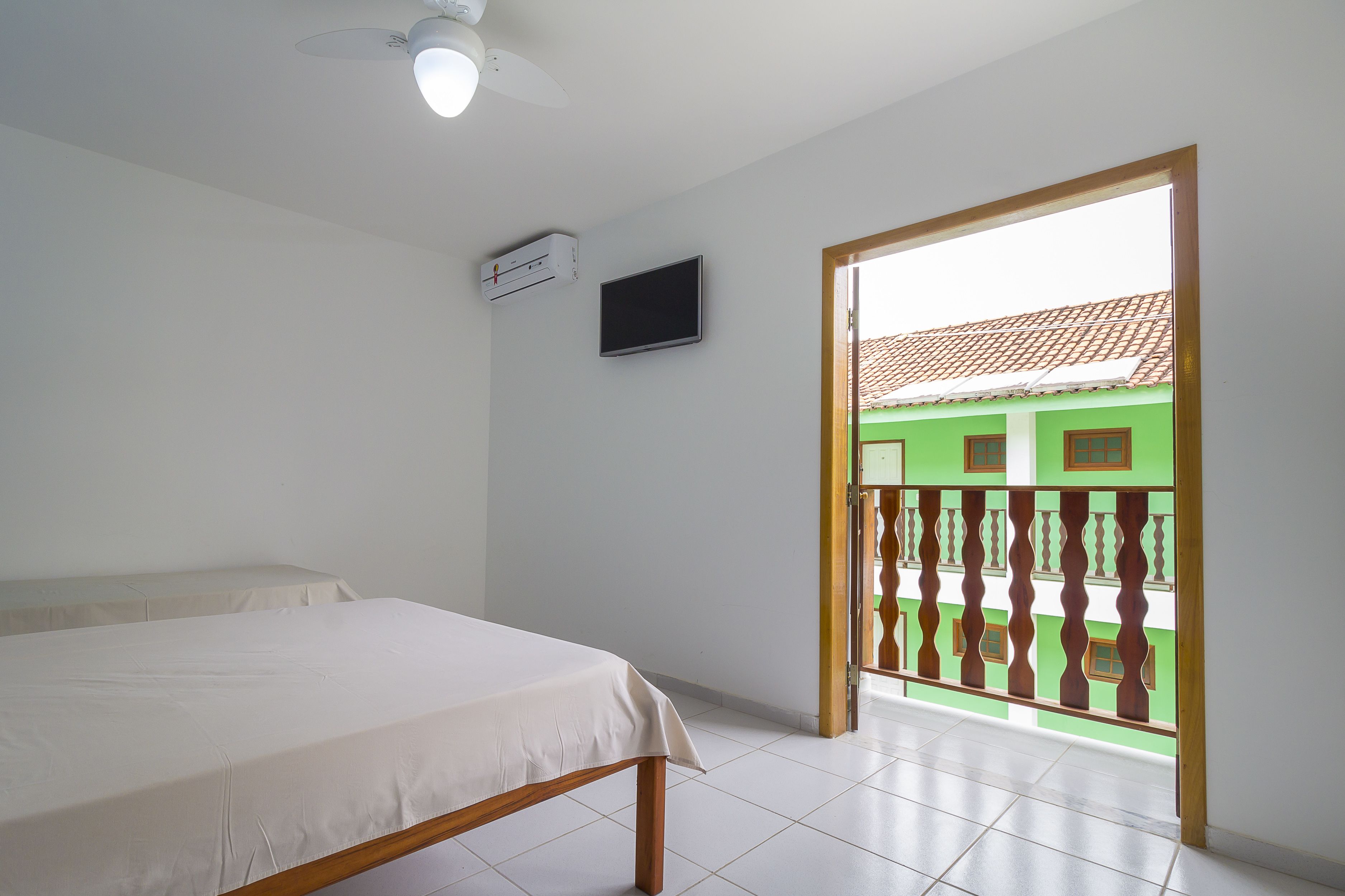 A hotel room with a bed, a ceiling fan, an air conditioning unit, a flat-screen TV, and a balcony with a view of a green building.
