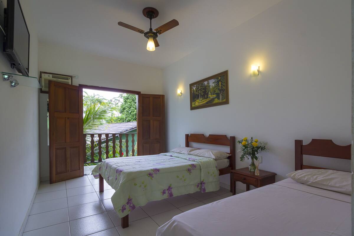 A bedroom with two beds, a ceiling fan, a nightstand with a vase of flowers, and a painting on the wall.