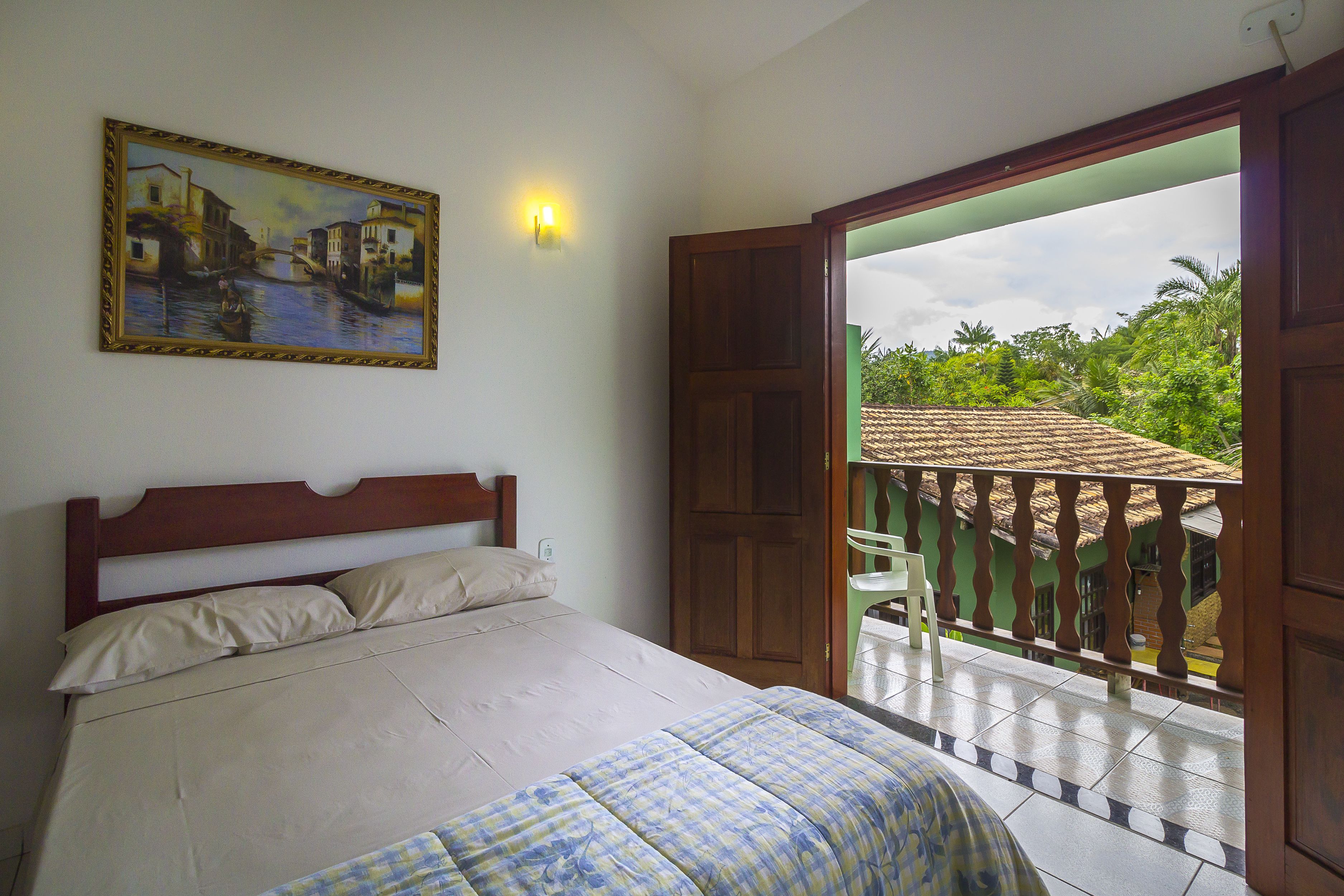 A bedroom with a bed, a painting, a nightstand, and a balcony with a view of greenery.