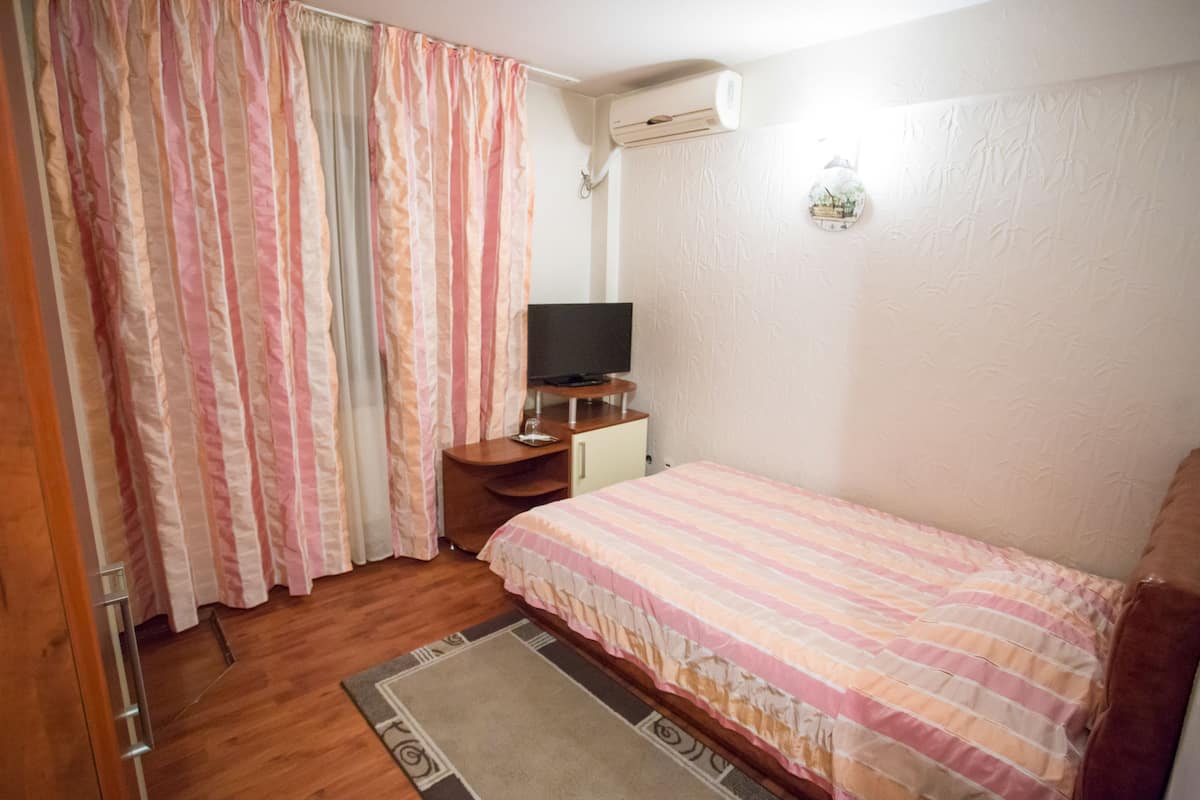 Quarto com cama, televisão sobre uma mesinha, ar-condicionado e cortina.