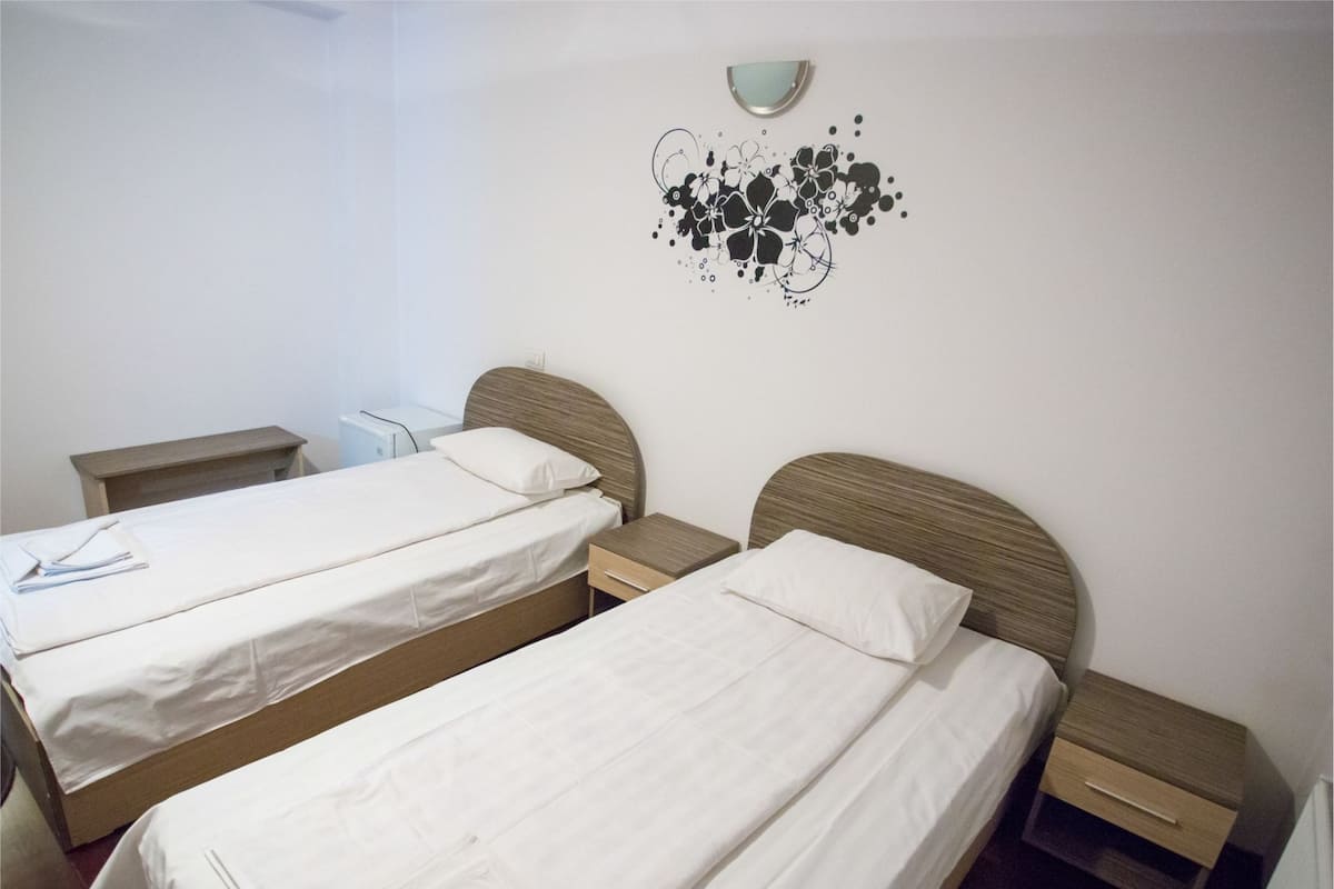 Quarto de hotel com duas camas de solteiro, cabeceiras de madeira, roupa de cama branca, mesas de cabeceira e uma decoração na parede.