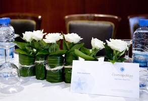 Una mesa con un cartel del Hotel Cosmopolitan, botellas de agua y rosas blancas en hojas verdes.