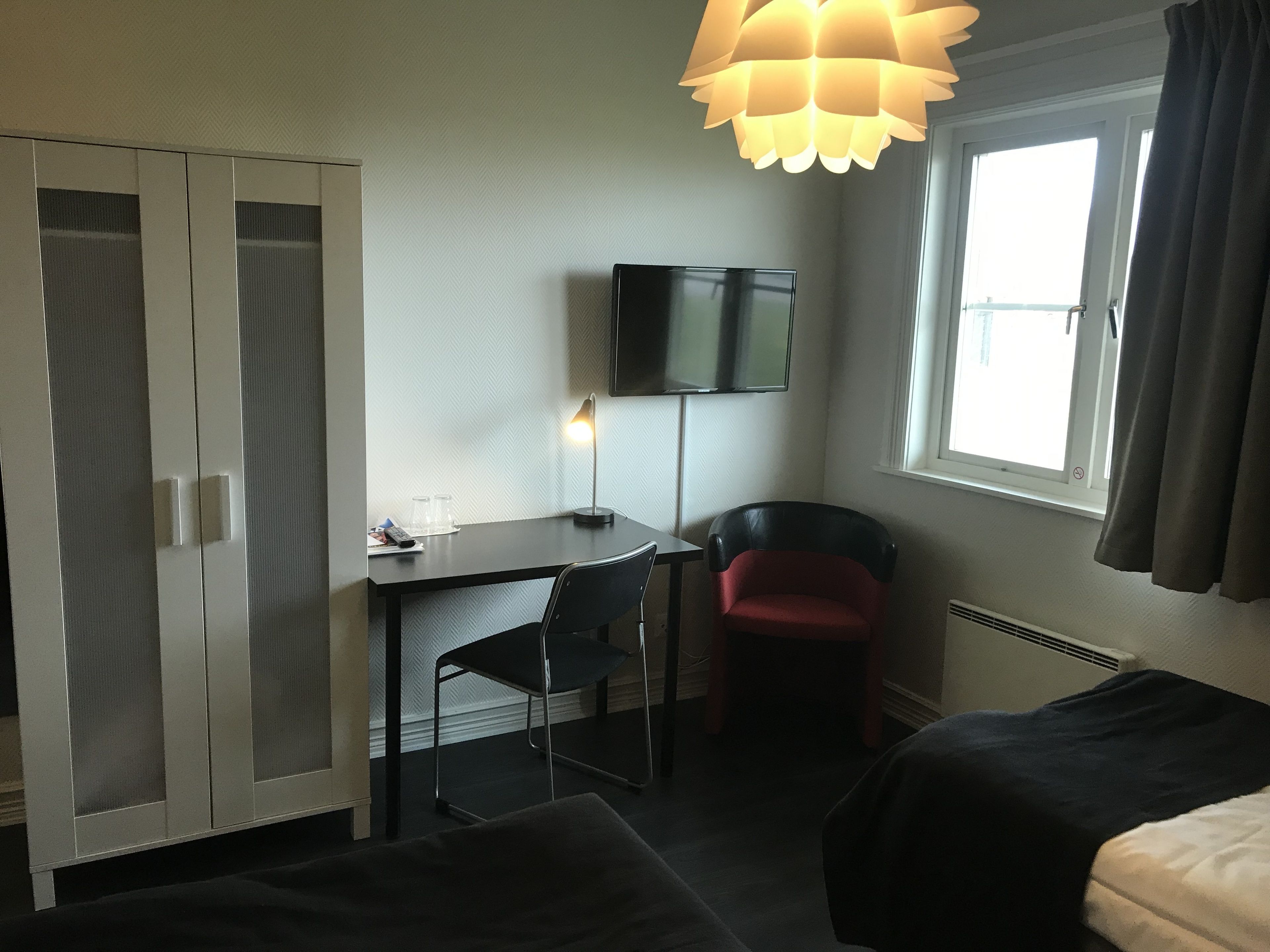 Habitación de hotel con cama, escritorio con silla, televisor montado en la pared y ventana con cortinas.