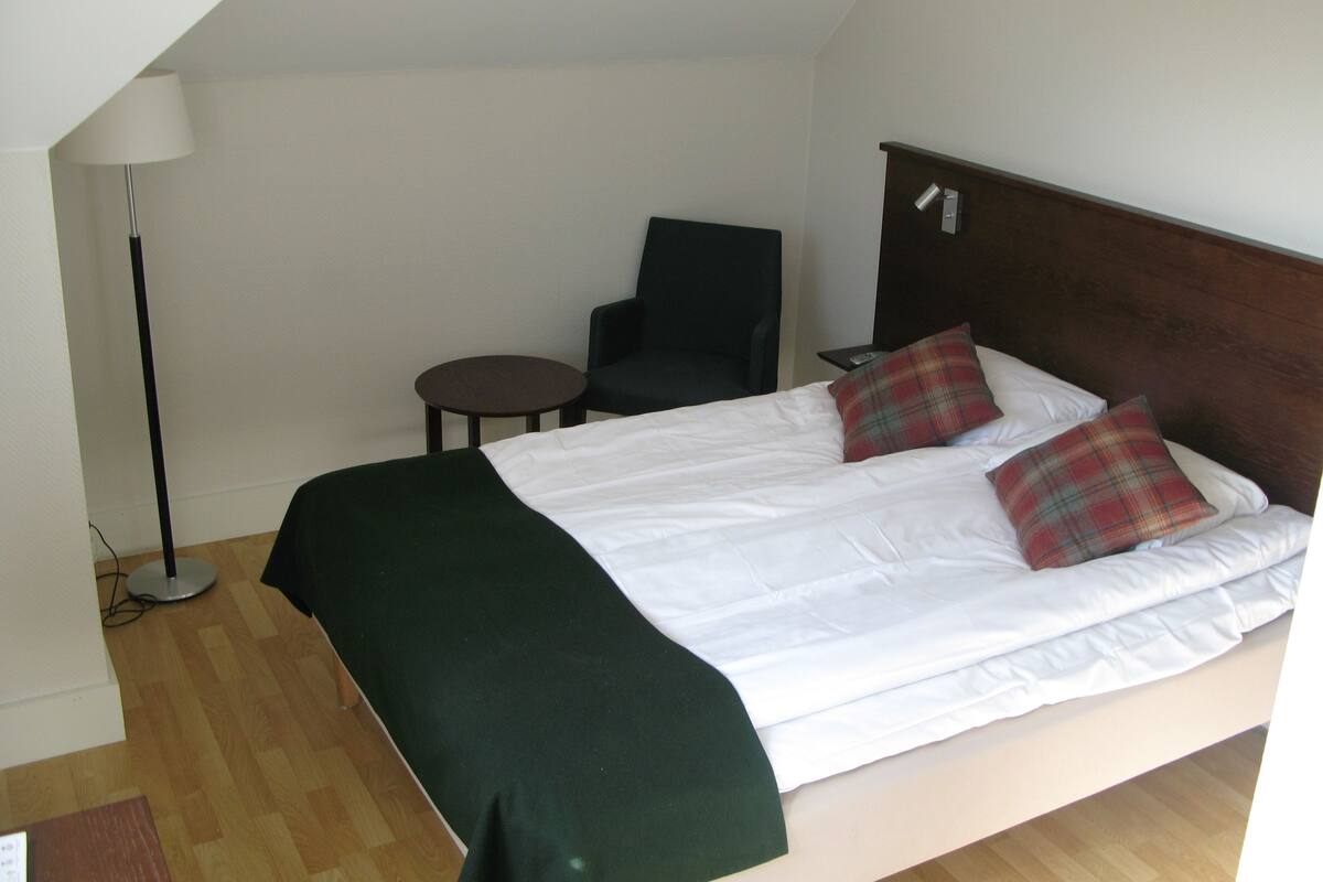 Una cama individual con dos almohadas, una mesita redonda pequeña, una silla y una lámpara de pie en una habitación con suelo de madera y paredes blancas.