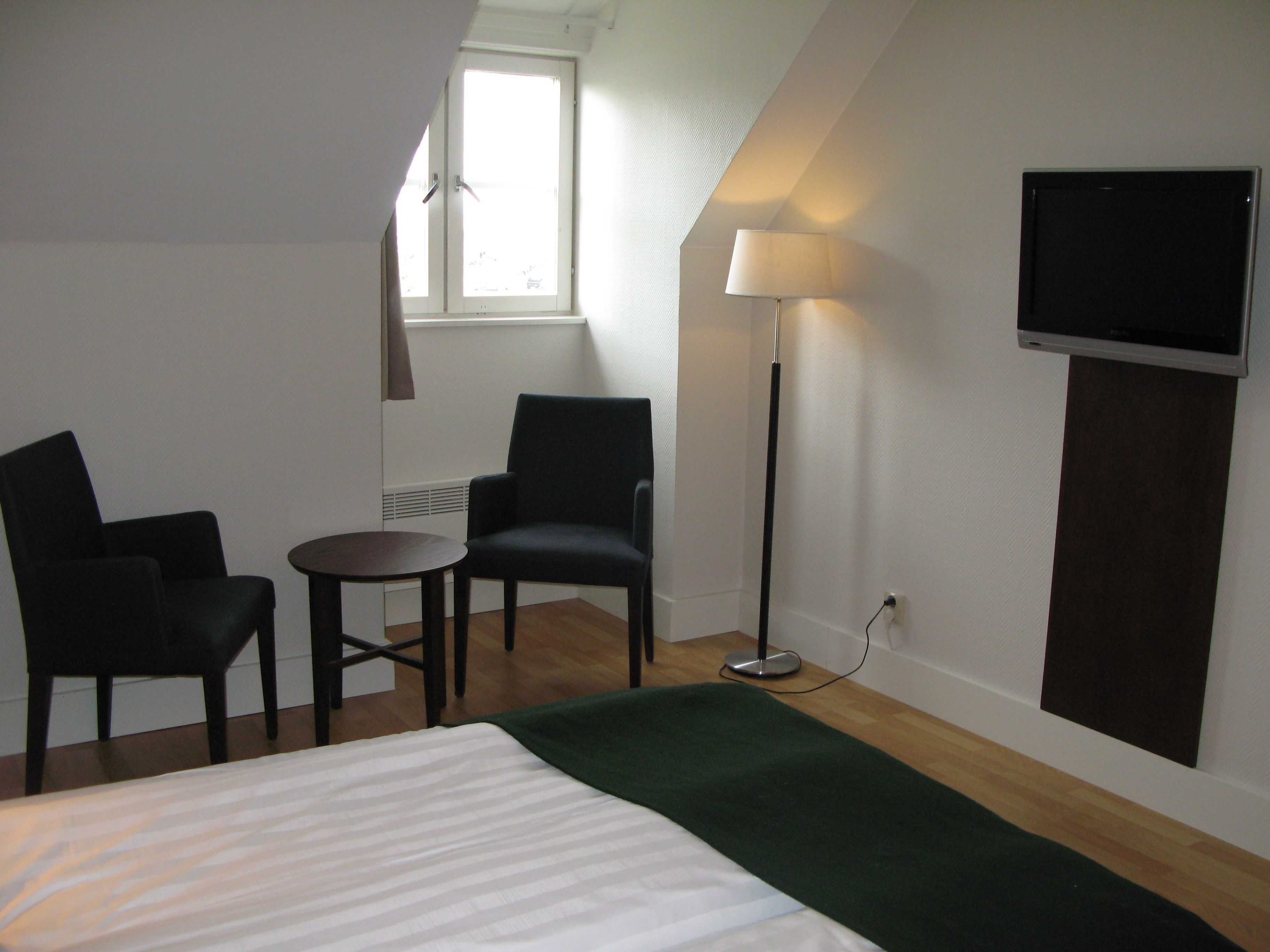 Una habitación de hotel con cama, dos sillas, una mesita, una lámpara de pie y televisor fijado a la pared.