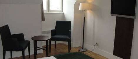 A hotel room with a bed, two chairs, a small table, a floor lamp, and a TV mounted on the wall.