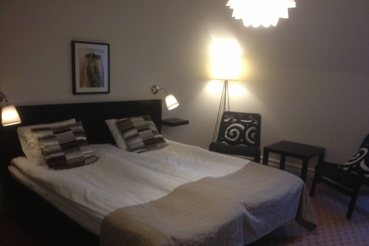 Una cama bien hecha con dos almohadas, una mesita de noche con lámpara, una silla e imagen enmarcada en la pared.