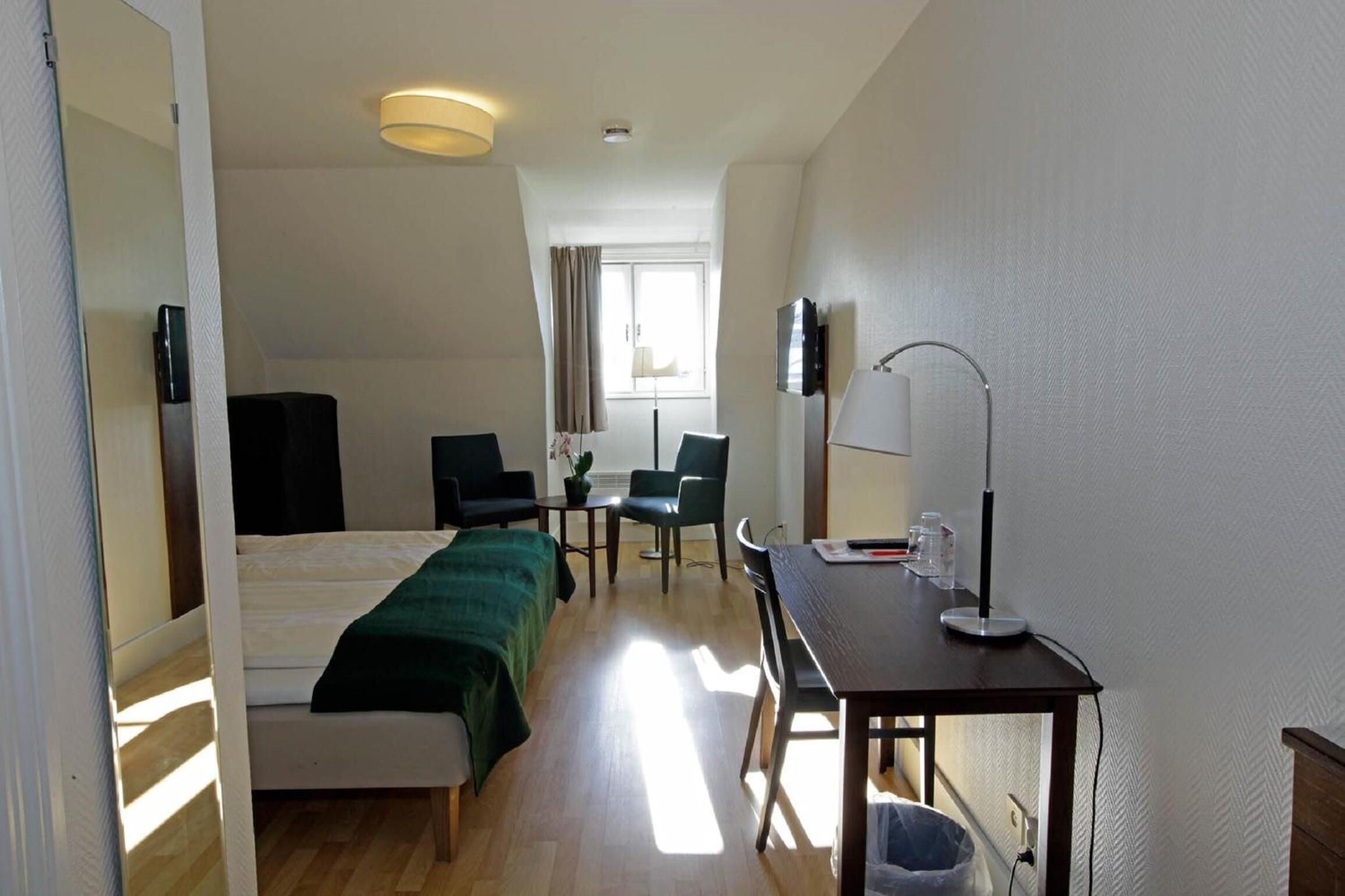 A hotel room with a bed, a desk with a lamp, a chair, a TV, and a window with curtains.