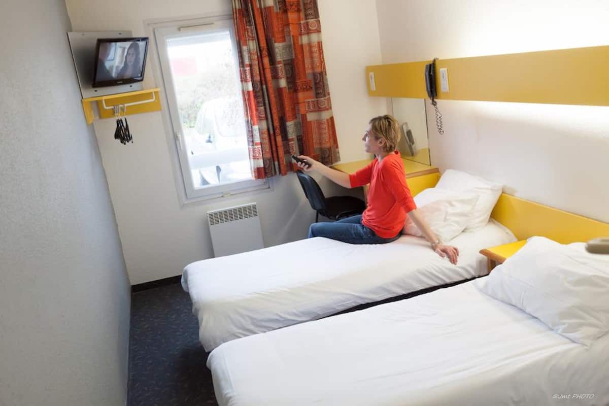 Une chambre d’hôtel avec deux lits simples, un téléviseur fixé au mur, un téléphone et une fenêtre avec des rideaux.
