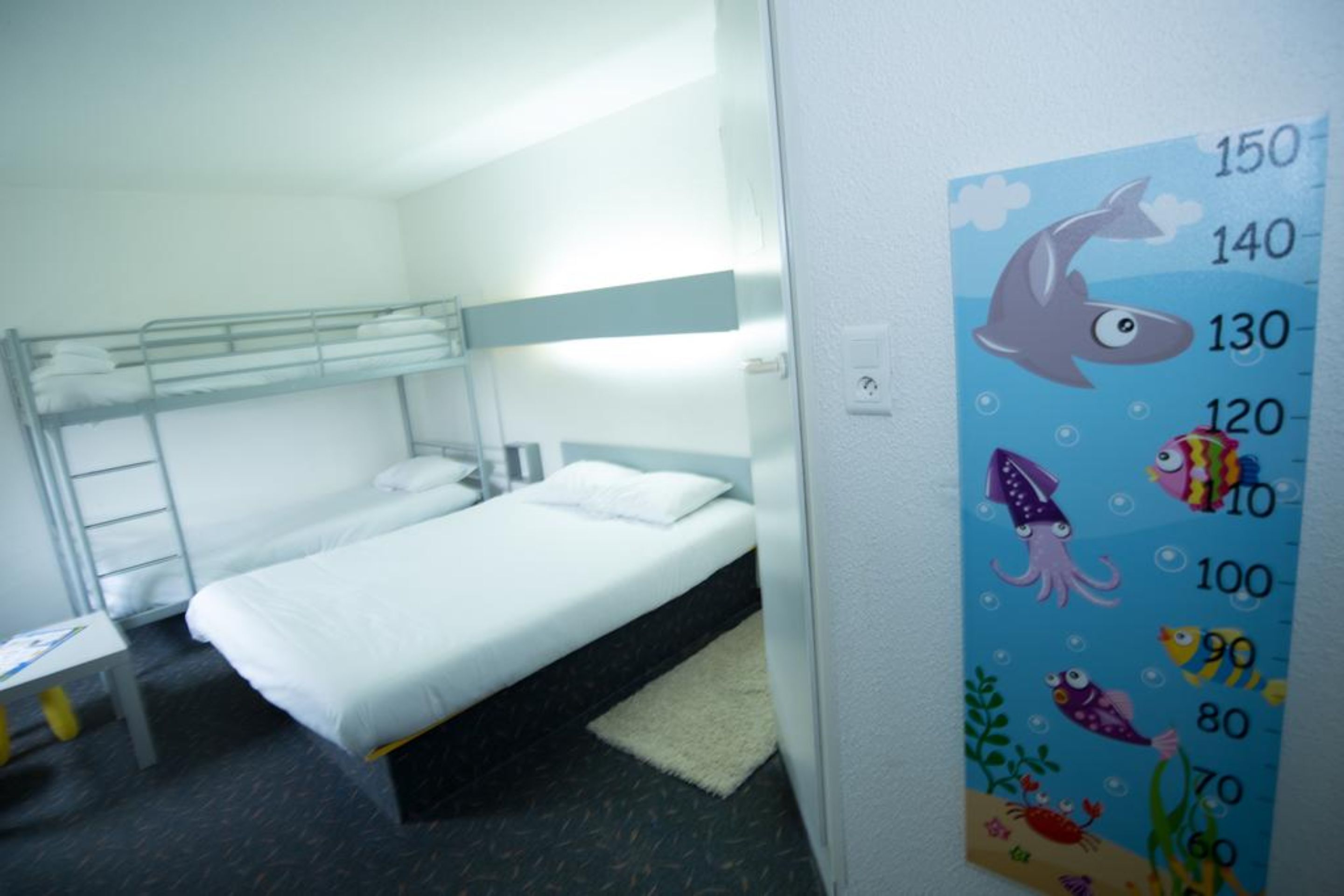 A single-bed room with a bunk bed above, a small table, and a wall with a height chart.
