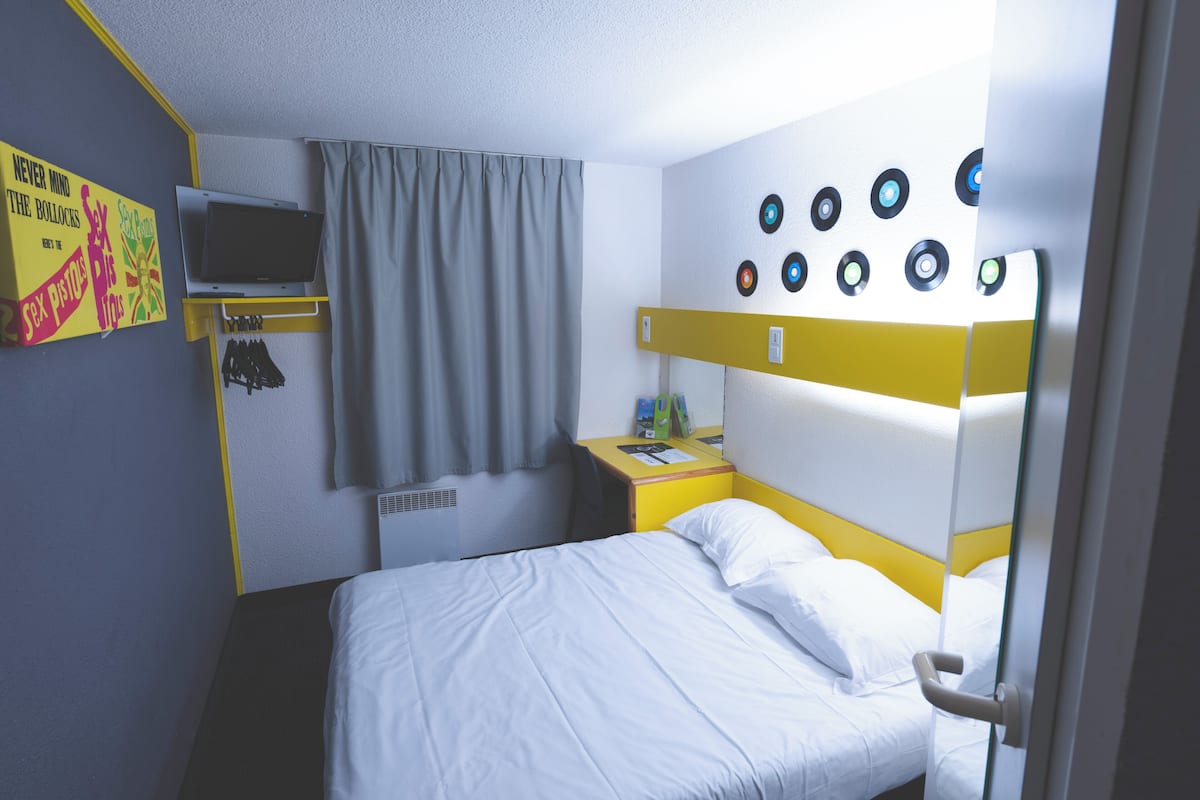 Une chambre d’hôtel avec un lit, une télévision, une table de chevet, une étagère fixée au mur avec des clés et un rideau.