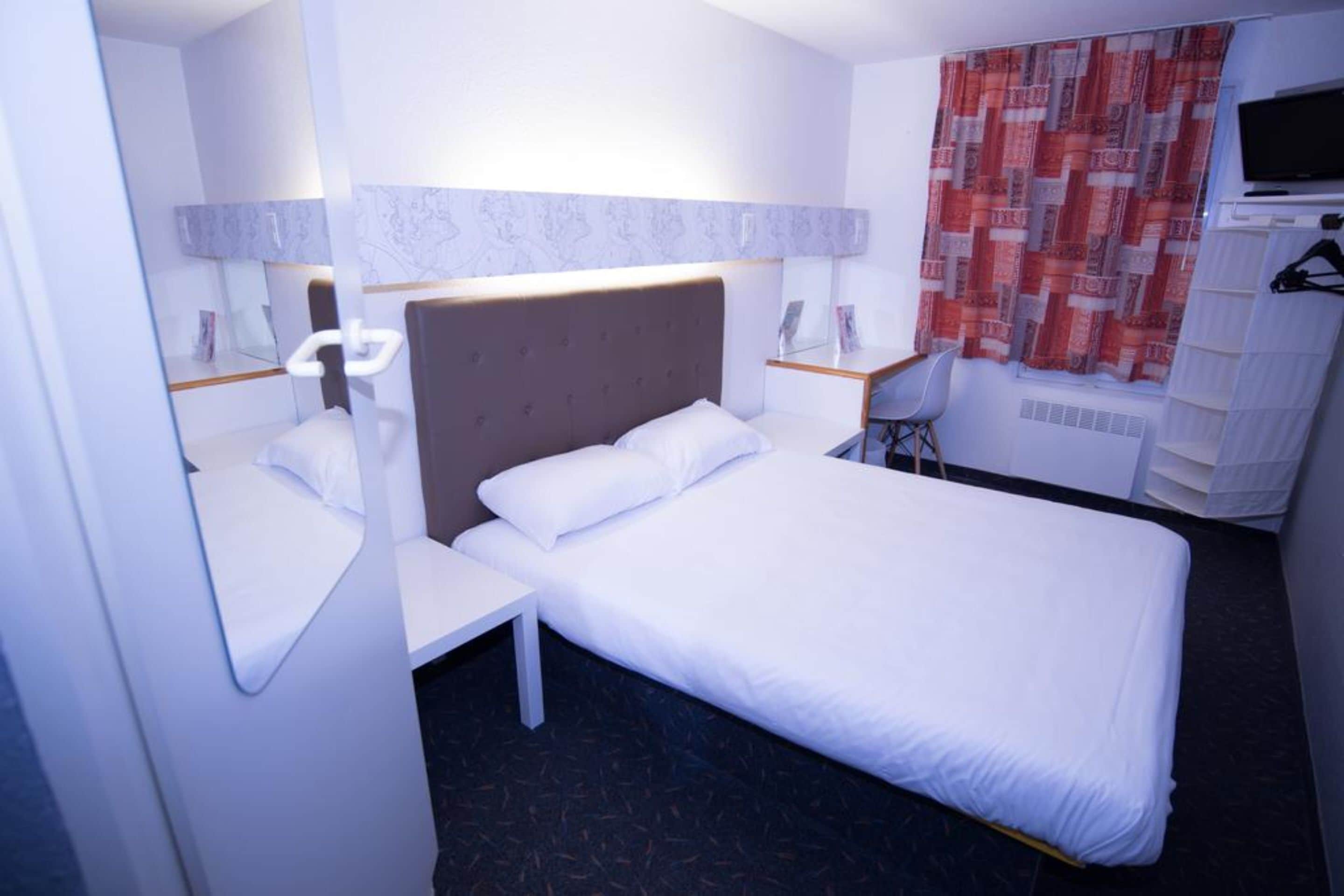 Habitación de hotel con cama, escritorio, silla, televisor y ventana con cortinas.