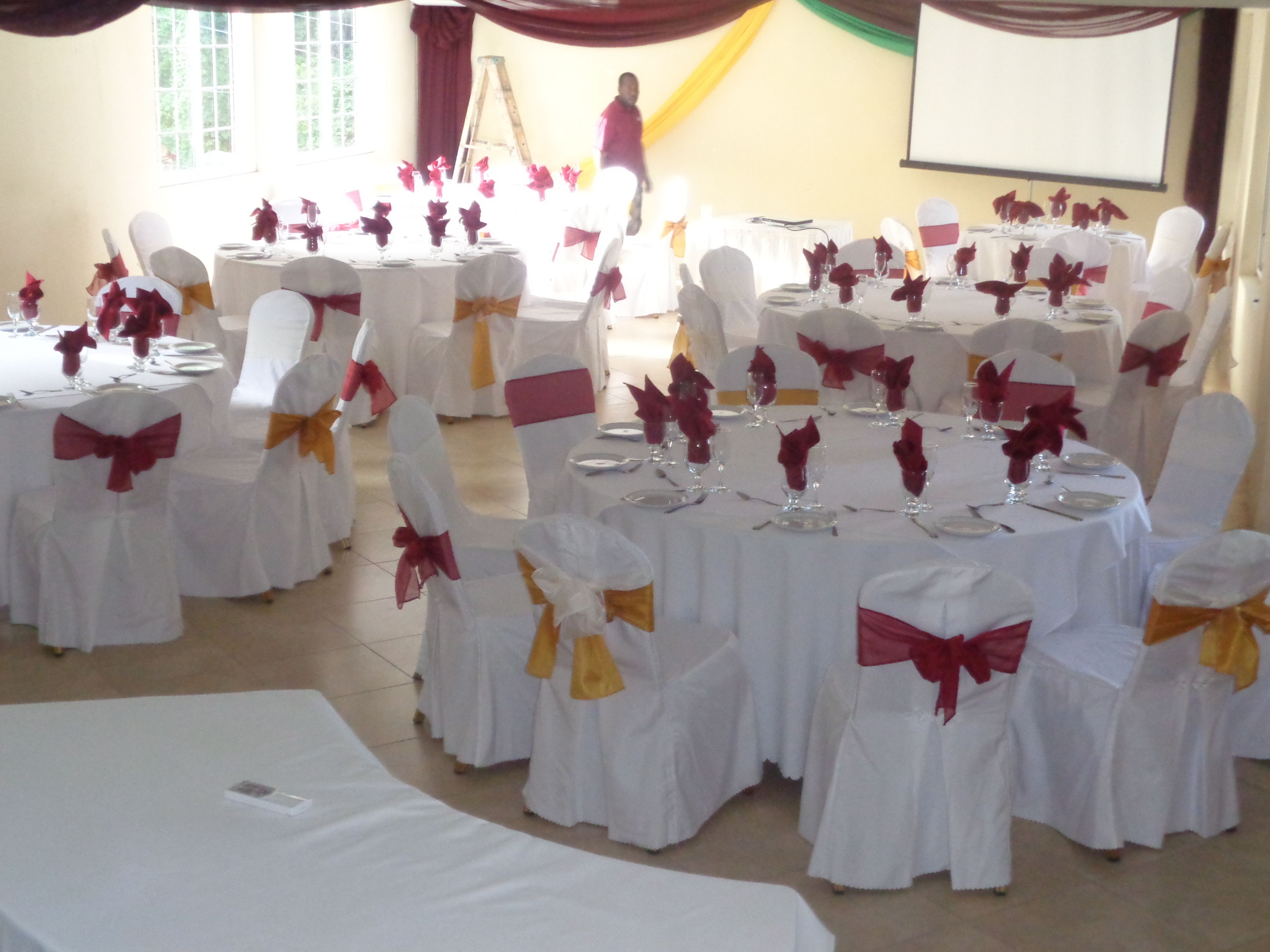 A banquet hall with round tables set for a formal event, each with white tablecloths and chairs adorned with red and yellow sashes.