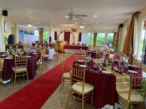 Formal dining area with round tables, red tablecloths, and gold chairs, set against a backdrop of lush greenery.