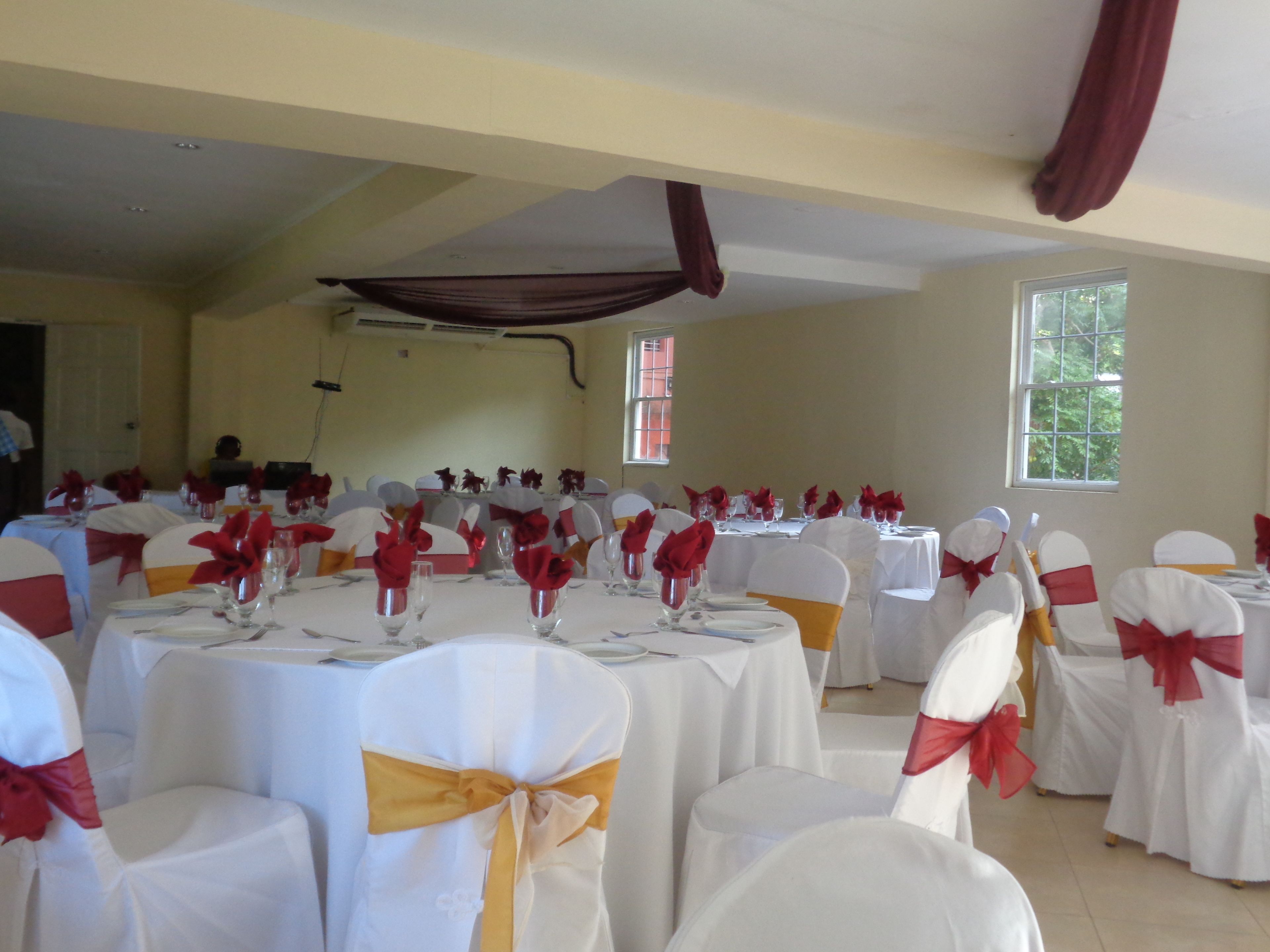 Un salón de banquetes con mesas redondas dispuestas para un evento formal, con manteles blancos y sillas adornadas con cintas rojas y amarillas.