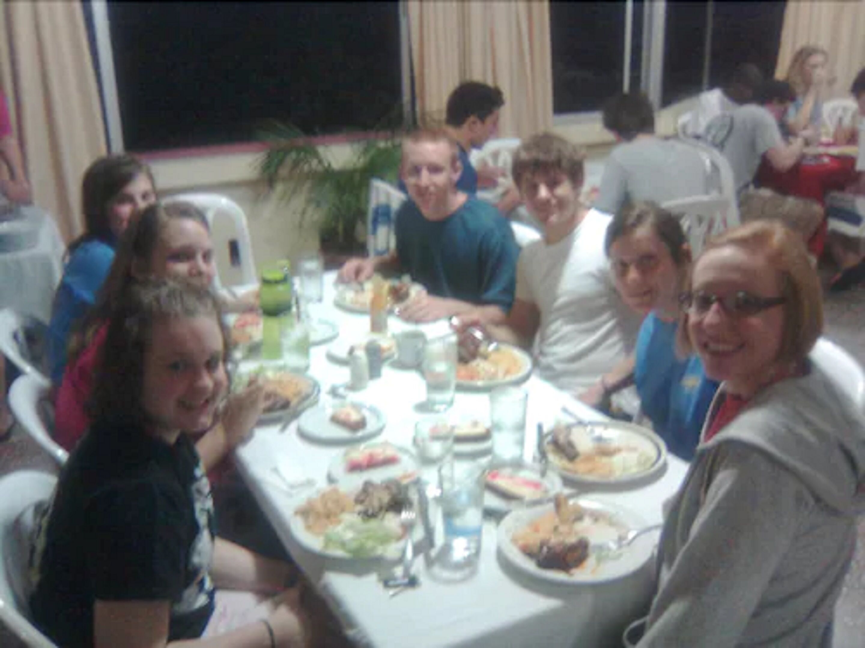A group of people seated around a table with various dishes, glasses, and a plant in the background.