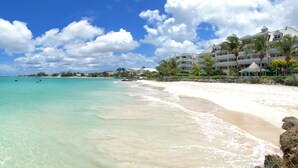 Una playa con agua turquesa cristalina, arena blanca y un resort con varias edificaciones y palmeras.