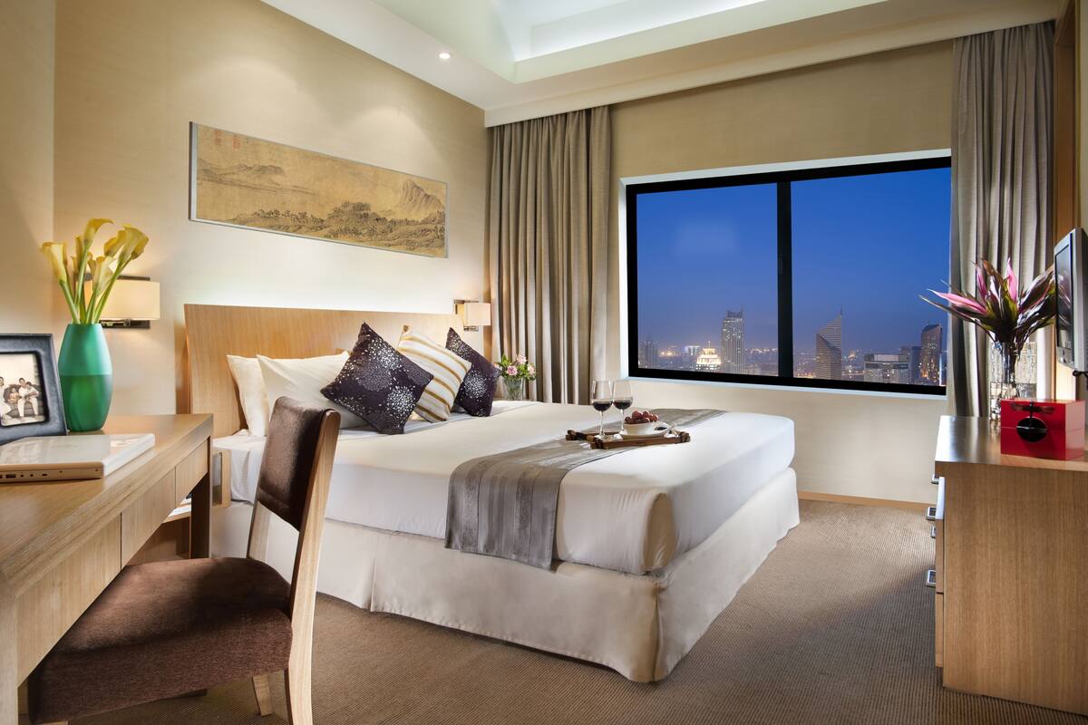 Une chambre d’hôtel avec un grand lit, un bureau avec un ordinateur portable, une chaise, une télévision et une vue sur la ville la nuit.