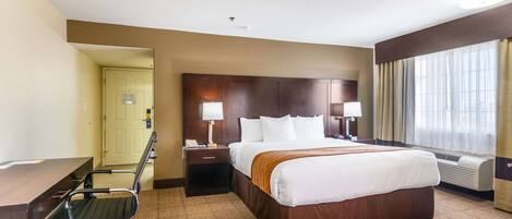 A hotel room with a large bed, a desk, a chair, a nightstand, and a window with curtains.
