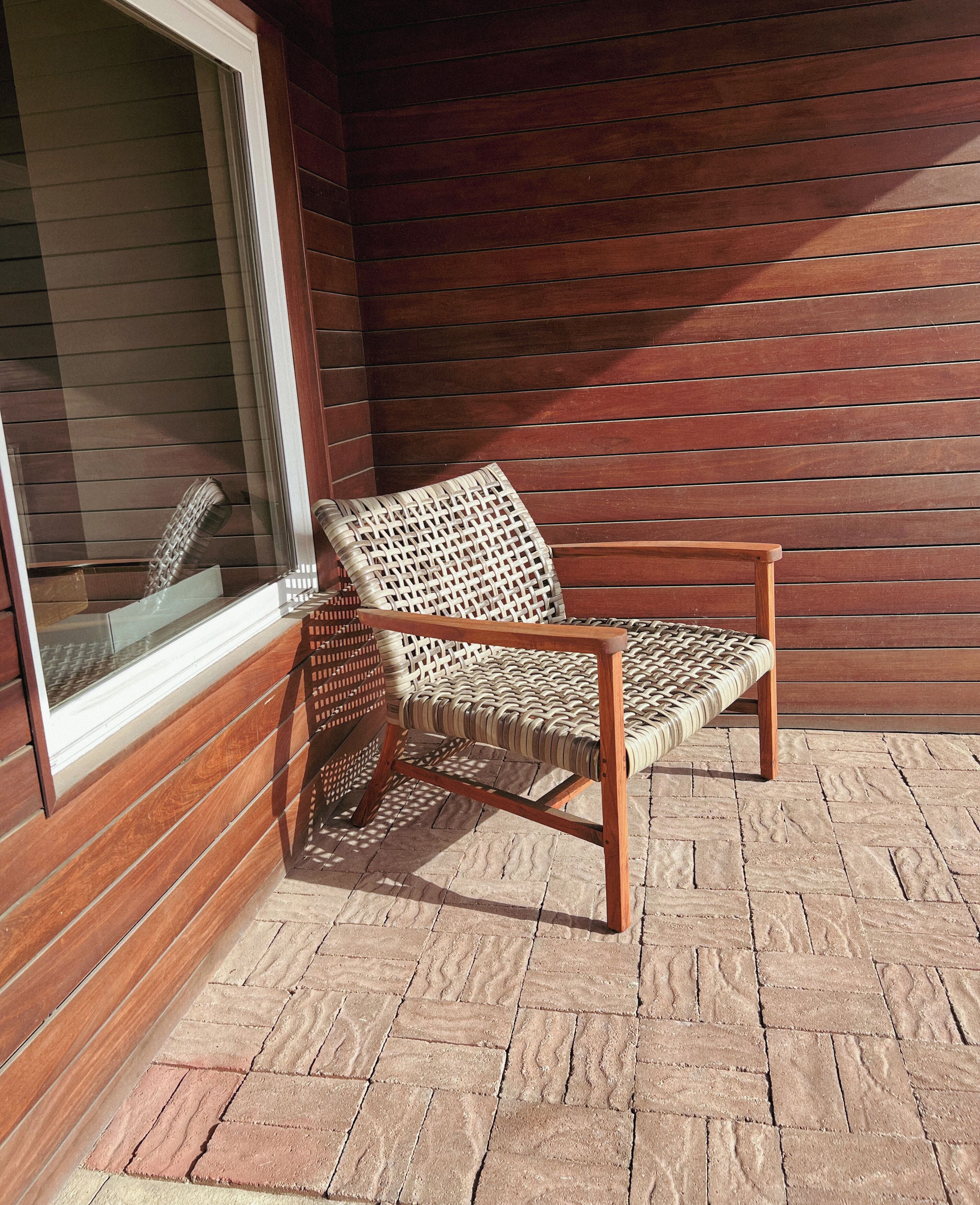 Una silla de mimbre con un cojín, ubicada en un patio de ladrillo junto a una pared de madera con revestimiento horizontal.