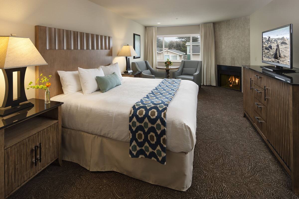 A hotel room with a large bed, two armchairs, a TV, a fireplace, and a view of the outside.