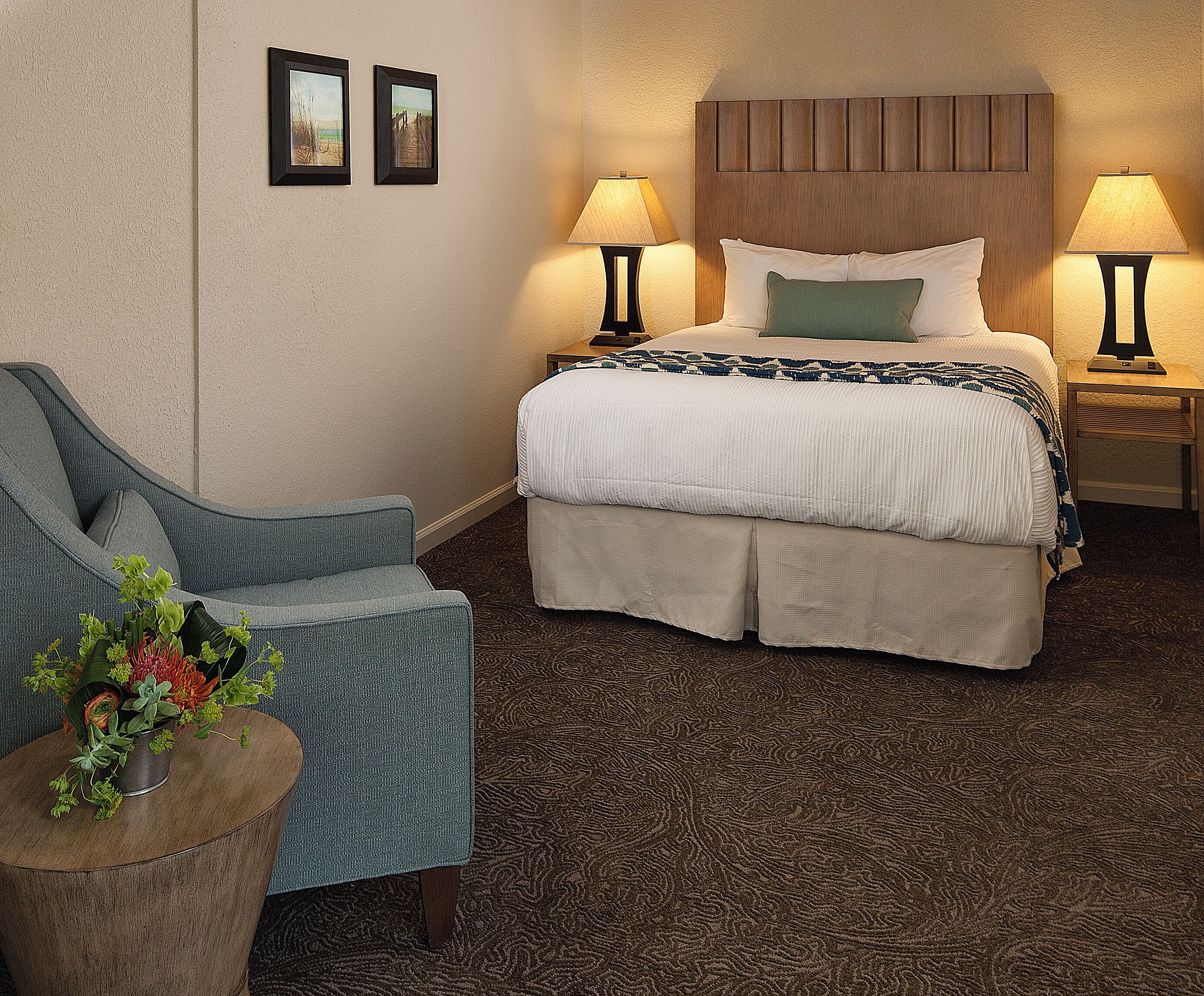 A hotel room with a bed, two bedside lamps, a chair, and a small table with a plant.