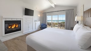 A hotel room with a large bed, a fireplace, a TV, and a balcony with a view of the ocean. - Cambria Landing Inn & Suites (Cambria)