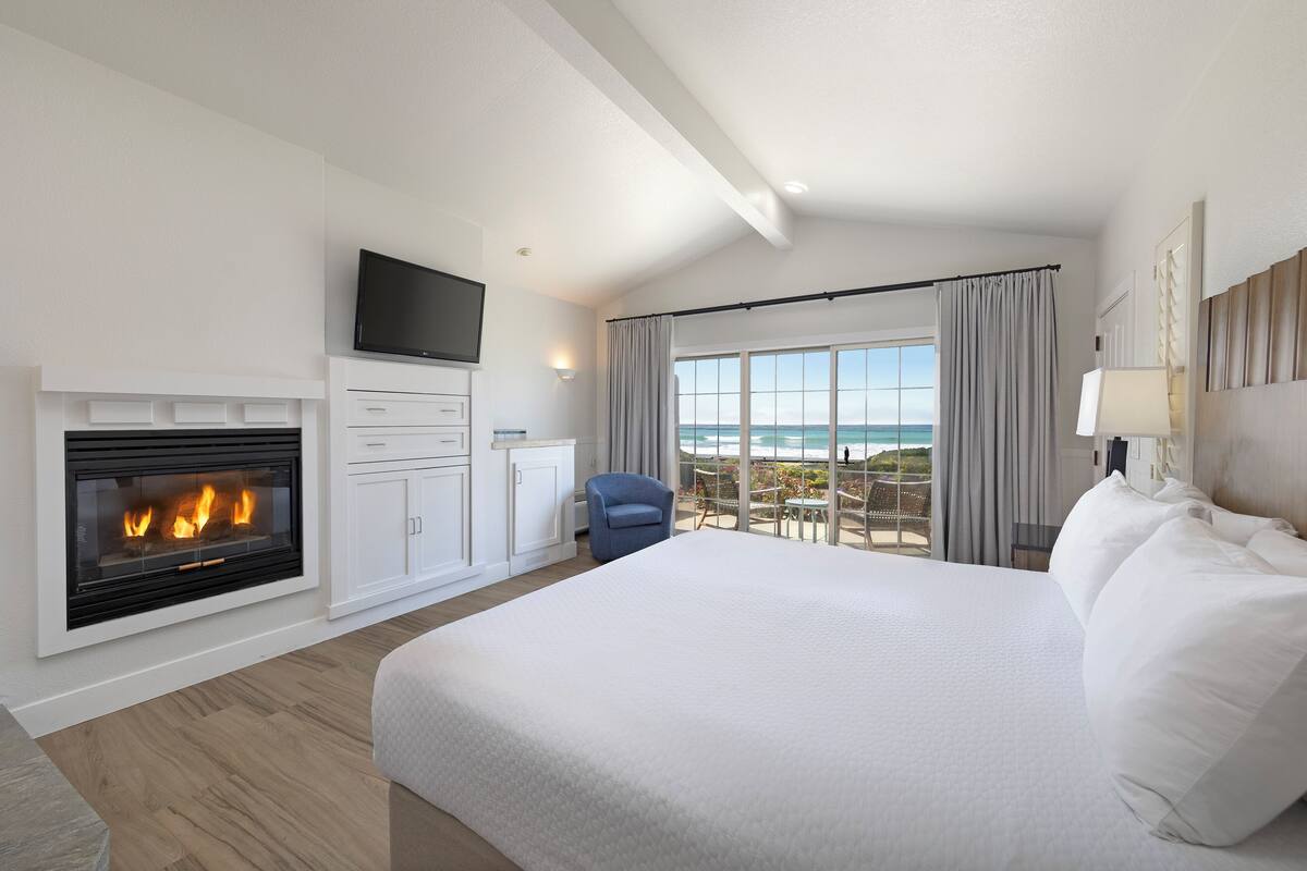 A hotel room with a large bed, a fireplace, a TV, and a balcony with a view of the ocean.