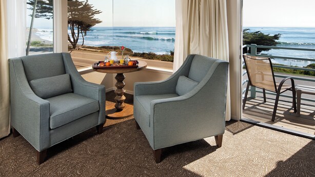 A room with two grey armchairs, a small round table with a tray of breakfast items, and a view of the ocean through large windows. - Cambria Landing Inn & Suites (Cambria)
