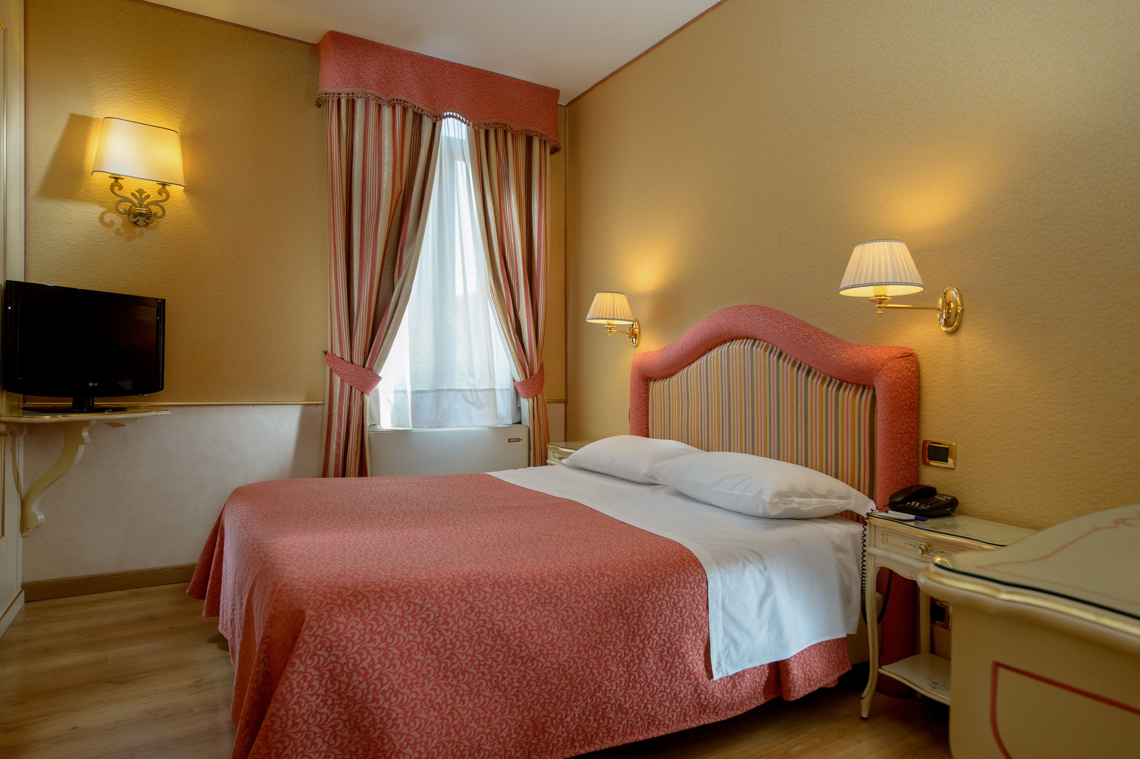 A hotel room with a large bed, a bedside table with a lamp, a television on a stand, and a window with curtains.