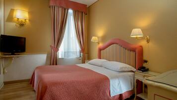 A hotel room with a large bed, a bedside table with a lamp, a television on a stand, and a window with curtains.